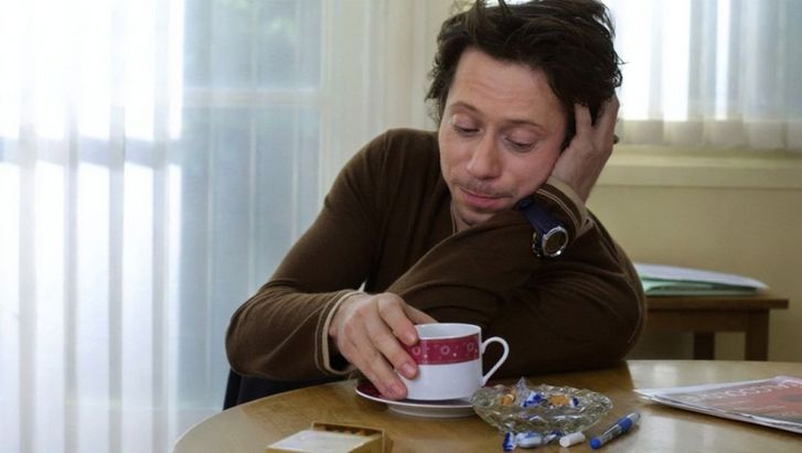 Film still from ROIS ET REINE. Mathieu Amalric is sitting at a table, a coffee cup in front of him.