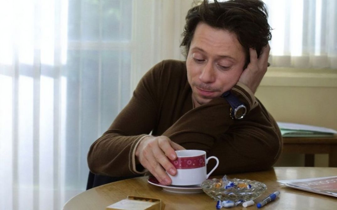 Filmstill aus ROIS ET REINE. Mathieu Amalric sitzt an einem Tisch, vor sich eine Kaffeetasse.