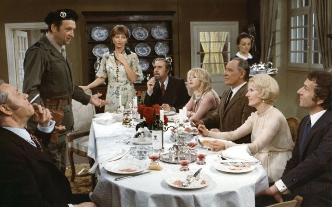 Film still from LE CHARME DISCRET DE LA BOURGEOISIE: A group of elegantly dressed people are sitting around a dining table.