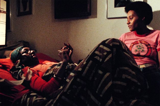 Film still from BORN IN FLAMES. Two women in a room talking, one lying, one sitting.