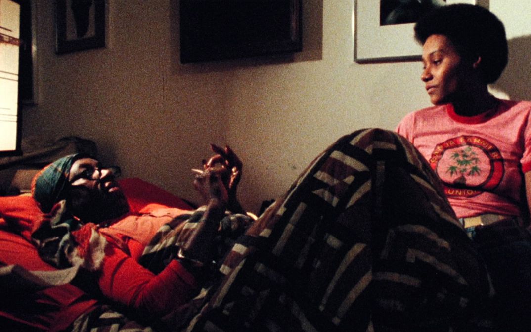 Film still from BORN IN FLAMES. Two women in a room talking, one lying, one sitting.