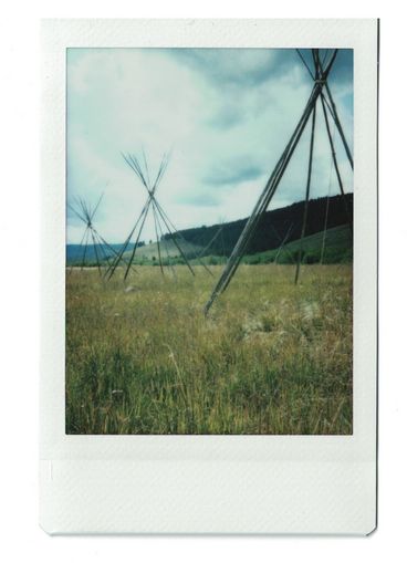 Polaroid of three tipi frames surrounded by greenish-brown grass in a field.