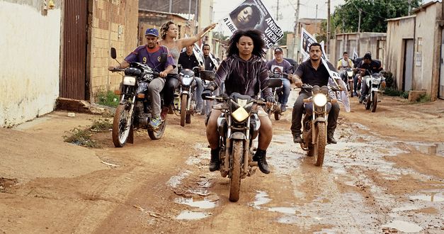 Filmstill aus „Mato seco em chamas (Dry Ground Burning)“ von Adirley Queirós und Joana Pimenta. Mehrere Menschen auf Motorrädern fahren in Formation durch eine Wohnsiedlung auf einer ungepflasterten Straßen. Eine Frau hält ein Transparent hoch. 