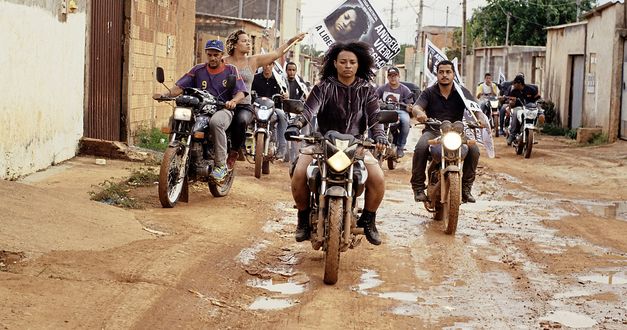 Still from the film "Mato seco em chamas (Dry Ground Burning)" by Adirley Queirós and Joana Pimenta. Several people on motorcycles ride in formation through a housing estate on an unpaved street. A woman holds up a banner.  