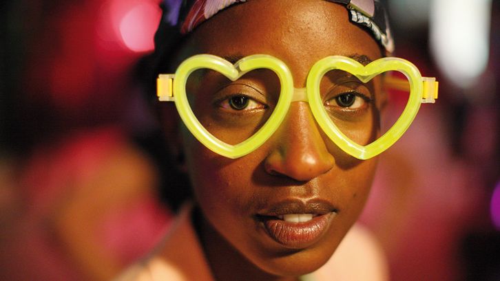 Film still from RAFIKI: A young woman wearing neon yellow glasses in the shape of a heart looks directly into the camera.