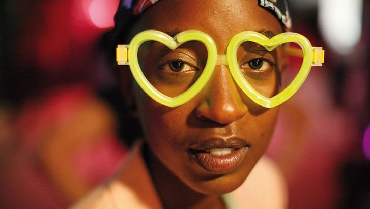 Film still from RAFIKI: A young woman wearing neon yellow glasses in the shape of a heart looks directly into the camera.