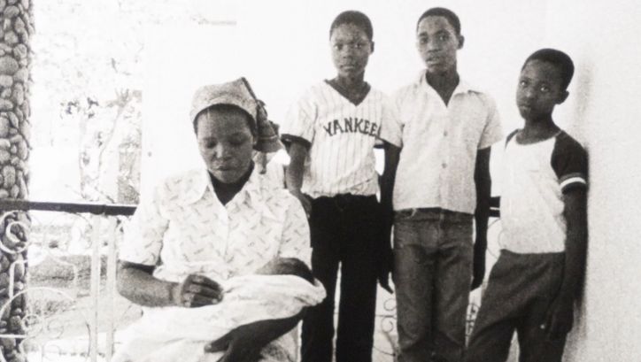 Film still from MANJACAZE. TEARS OF OUR HOPE: A woman sits on a porch with a baby on her lap. Behind her are three boys.