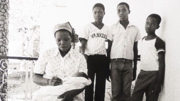 Film still from MANJACAZE. TEARS OF OUR HOPE: A woman sits on a porch with a baby on her lap. Behind her are three boys.