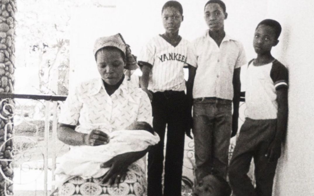 Film still from MANJACAZE. TEARS OF OUR HOPE: A woman sits on a porch with a baby on her lap. Behind her are three boys.