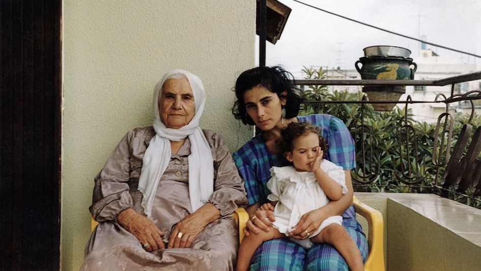 Film still from BYE BYE TIBERIAS: An old and a young woman are sitting next to each other on a balcony. The younger woman has a little girl on her lap.