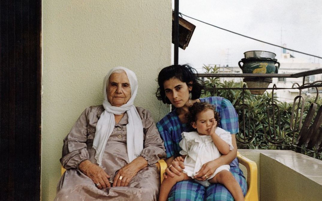 Filmstill aus BYE BYE TIBERIAS: Eine alte und eine junge Frau sitzen nebeneinander auf einem Balkon. Die jüngere Frau hat ein kleines Mädchen auf dem Schoß.