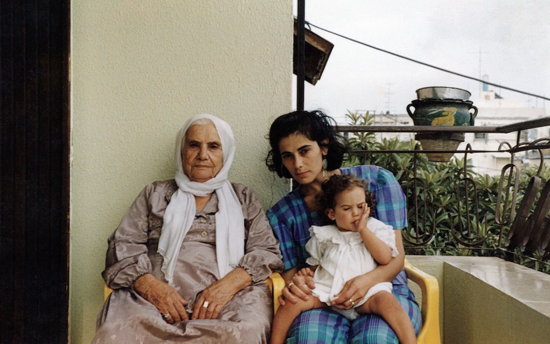 Filmstill aus BYE BYE TIBERIAS: Eine alte und eine junge Frau sitzen nebeneinander auf einem Balkon. Die jüngere Frau hat ein kleines Mädchen auf dem Schoß.
