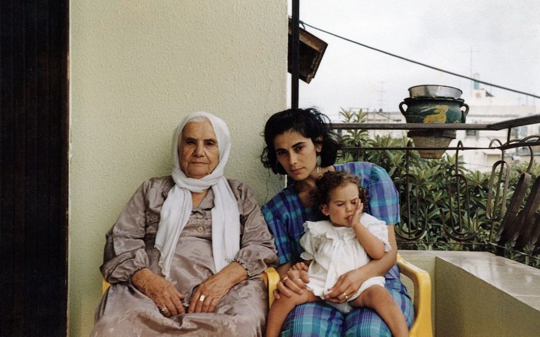 Film still from BYE BYE TIBERIAS: An old and a young woman are sitting next to each other on a balcony. The younger woman has a little girl on her lap.