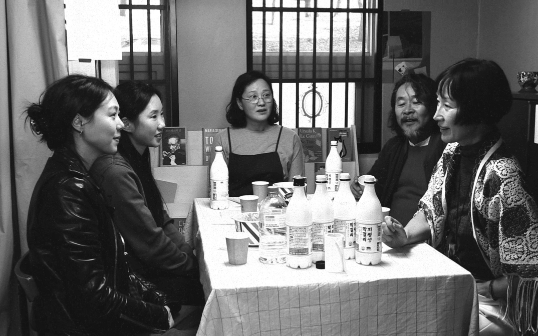 Filmstill aus Die Schriftstellerin, ihr Film und ein glücklicher Zufall: Fünf Menschen sitzen rund um einen Tisch, der voller alkoholischer Getränke ist.