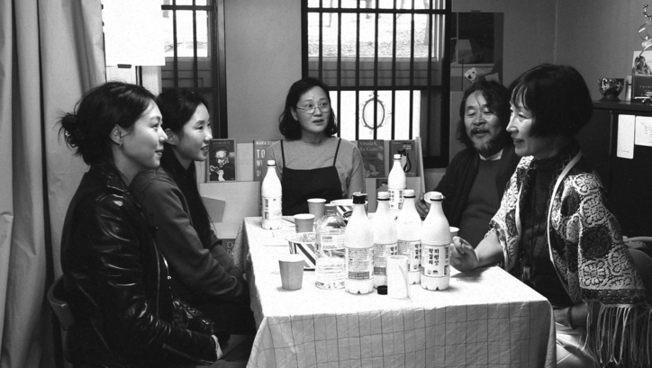 Filmstill aus Die Schriftstellerin, ihr Film und ein glücklicher Zufall: Fünf Menschen sitzen rund um einen Tisch, der voller alkoholischer Getränke ist.