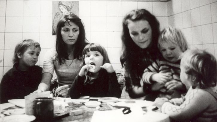 Film still from GELEGENHEITSARBEIT EINER SKLAVIN. Two women and four children sit at a table.