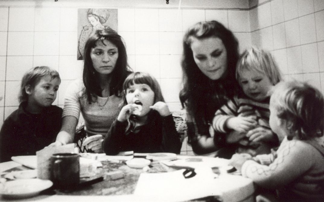 Film still from GELEGENHEITSARBEIT EINER SKLAVIN. Two women and four children sit at a table.