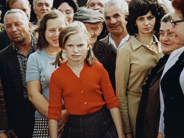 Film still from Zuza Banasińska’s film “Grandmamauntsistercat”. A young girl with pigtails in a bright red cardigan stands with a crowd of older people. It is unclear what they are observing behind the camera. The image is distorted, creating a noticeably odd and unconventional representation of the girl’s face. 