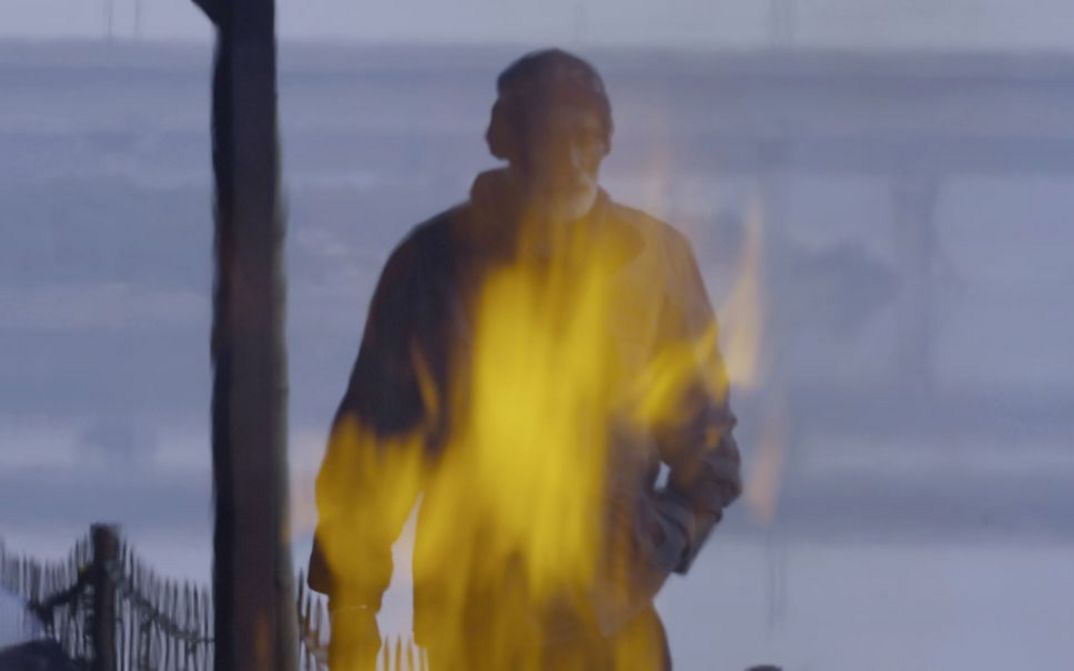 Still from the film "The Wind in Your Body Is Just Visiting, Your Breath Will Soon Be Thunder" by Pallavi Paul. A fire burns in the foreground, in the background a man walks towards the camera.