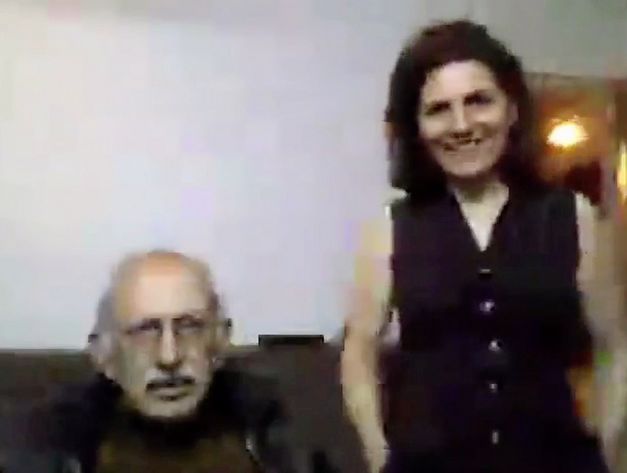 Film still from "Was hast du gestern geträumt, Parajanov?" by Faraz Fesharaki. It shows a blurred close-up of an older man sitting and a woman standing to his right. They are looking towards the camera.