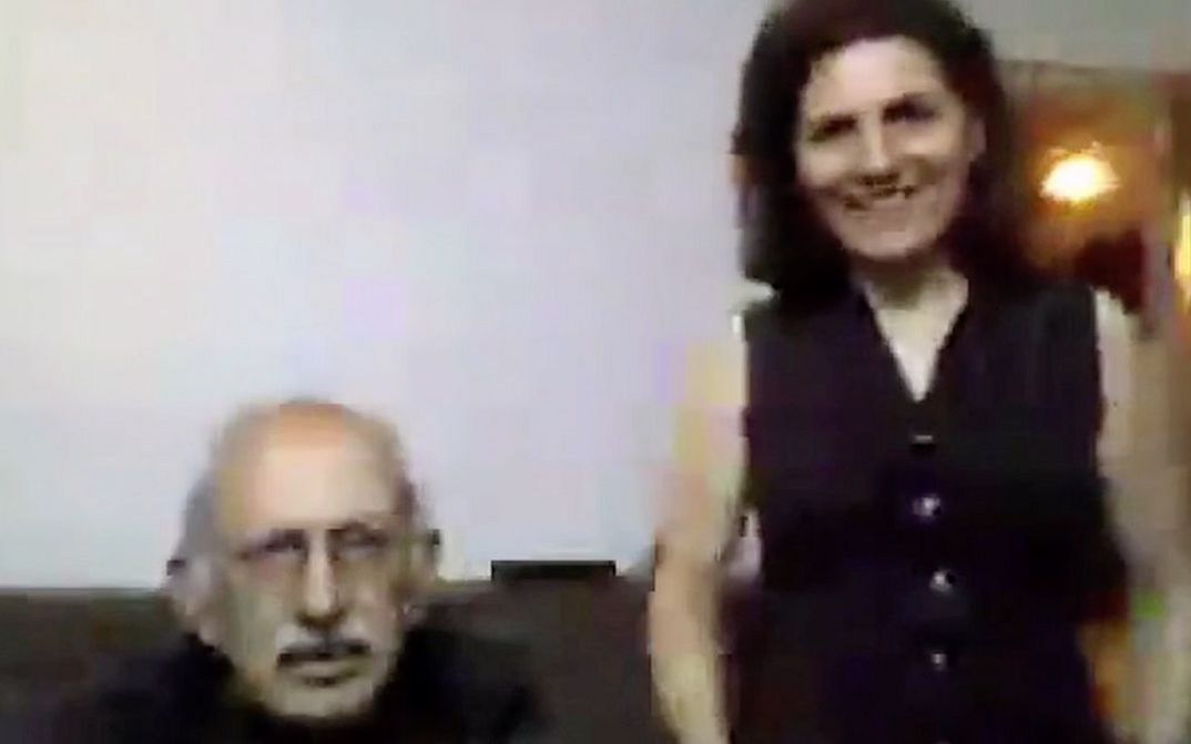 Filmstill aus "Was hast du gestern geträumt, Parajanov?" von Faraz Fesharaki. Zu sehen ist eine unscharfe Nahaufnahme eines älteren Mannes, der sitzt, und einer Frau, die rechts von ihm steht. Sie schauen in Richtung Kamera.