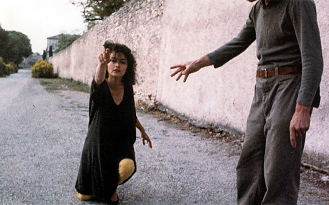 Filmstill aus NEUER ENGEL. WESTWÄRTS. Eine Frau kniet auf einer Straße und zeigt auf etwas außerhalb des Bildes.