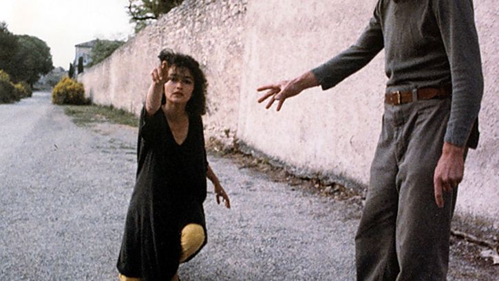 Film still from NEUER ENGEL. WESTWÄRTS. A woman kneels on a street and points to something outside the picture.