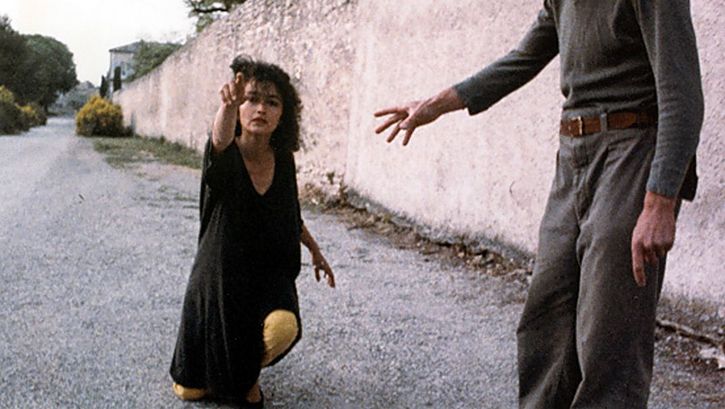 Filmstill aus NEUER ENGEL. WESTWÄRTS. Eine Frau kniet auf einer Straße und zeigt auf etwas außerhalb des Bildes.