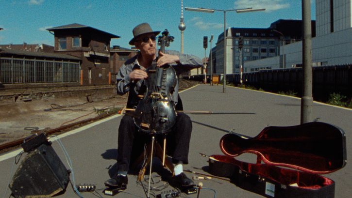 Film still from BERLIN - BAHNHOF FRIEDRICHSTRASSE: A man plays the cello and sits on a street. Next to it is an old railway station building and the Berlin television tower in the background.