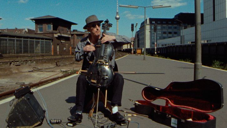 Film still from BERLIN - BAHNHOF FRIEDRICHSTRASSE: A man plays the cello and sits on a street. Next to it is an old railway station building and the Berlin television tower in the background.