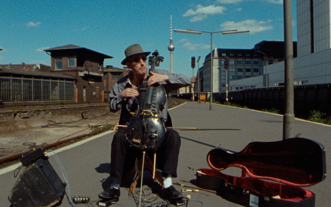 Filmstill aus BERLIN - BAHNHOF FRIEDRICHSTRASSE: Ein Mann spielt Cello und sitzt auf einer Straße. Daneben ein altes Bahnhofsgebäude und im Hintergrund der Berliner Fernsehrturm.