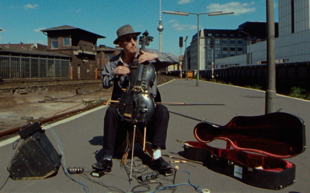 Filmstill aus BERLIN - BAHNHOF FRIEDRICHSTRASSE: Ein Mann spielt Cello und sitzt auf einer Straße. Daneben ein altes Bahnhofsgebäude und im Hintergrund der Berliner Fernsehrturm.