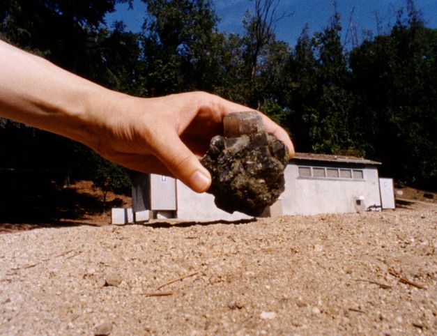 Filmstill aus dem Film "Instant Life" von Anja Dornieden, Juan David González Monroy und Andrew Kim. Man sieht eine Hand einen Kristall halten, im Hintergrund steht ein Haus.