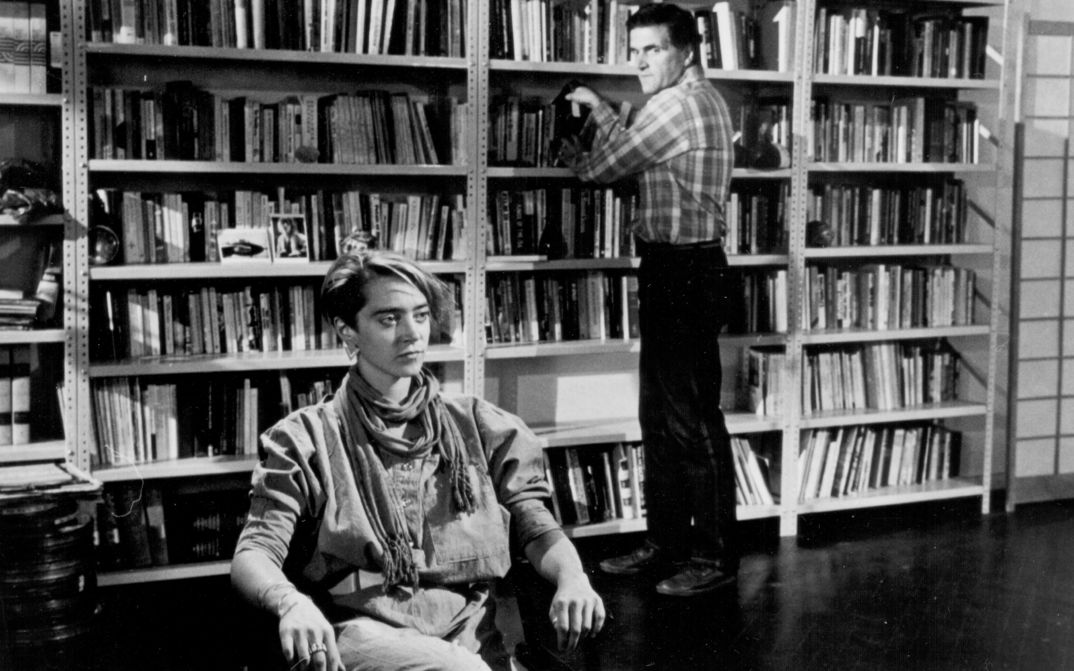 Filmstill aus THE MAN WHO ENVIED WOMEN: Ein Mann steht an einem Bücherregal, davor sitzt eine Frau.