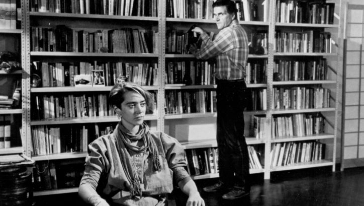 Film still from THE MAN WHO ENVIED WOMEN: A man stands at a bookshelf, a woman sits in front of it.