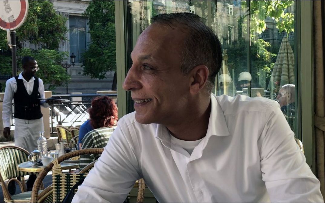 Mann sitzt an einem Cafétisch im Freien und blickt zur Seite, Kellner und Gäste im Hintergrund.