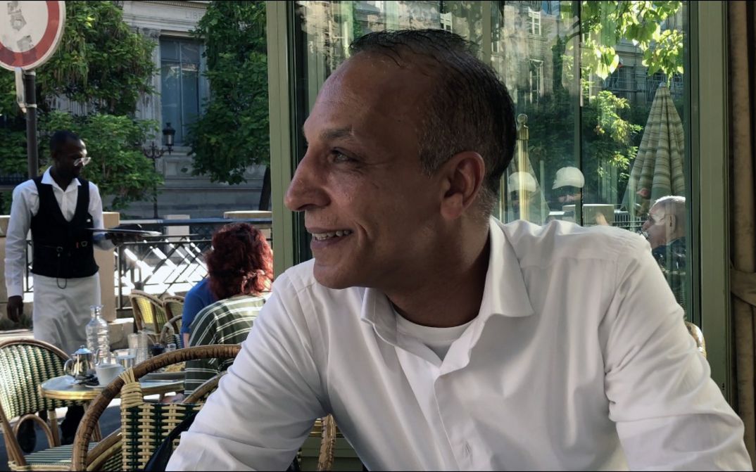 Mann sitzt an einem Cafétisch im Freien und blickt zur Seite, Kellner und Gäste im Hintergrund.
