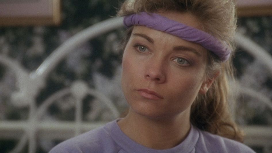 Filmstill aus TRACK 29: Eine Frau mit lila Sweatshirt und Stirnband schaut nachdenklich auf einen Fernseher außerhalb des Bildes.