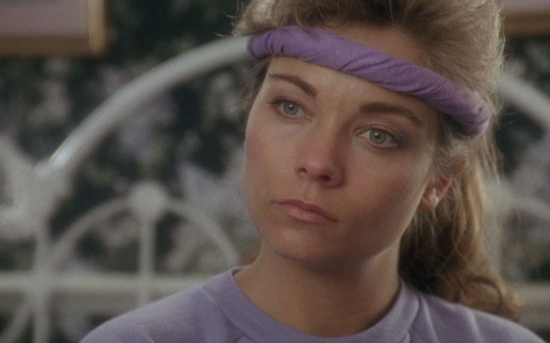 Filmstill aus TRACK 29: Eine Frau mit lila Sweatshirt und Stirnband schaut nachdenklich auf einen Fernseher außerhalb des Bildes.