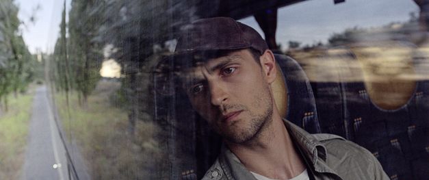 Film still from "Redaktsiya" by Roman Bondarchuk. It shows a close-up of a man in a cap looking through the window of a moving vehicle. 