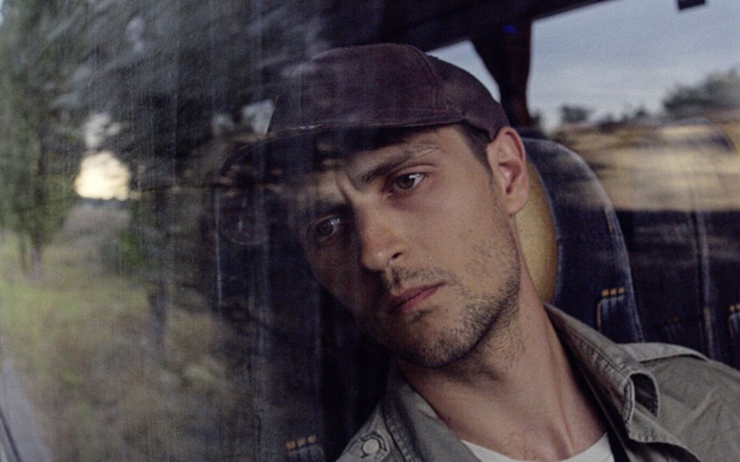 Film still from "Redaktsiya" by Roman Bondarchuk. It shows a close-up of a man in a cap looking through the window of a moving vehicle. 