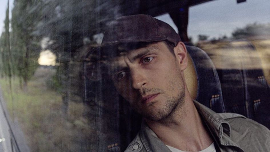Film still from "Redaktsiya" by Roman Bondarchuk. It shows a close-up of a man in a cap looking through the window of a moving vehicle. 