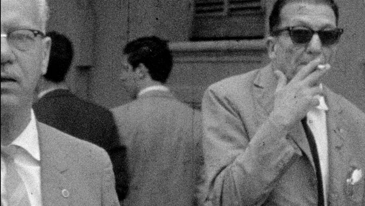 Filmstill aus BAJO LAS BANDERAS, EL SOL: Ein verschwommenes Fernseh-Bild in schwarz-weiß. Man sieht zwei ältere Männer in Anzug auf der Straße, einer raucht eine Zigarette. Im Hintergrund sind weitere Männer.