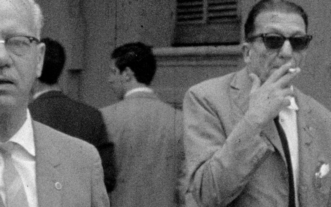 Filmstill aus BAJO LAS BANDERAS, EL SOL: Ein verschwommenes Fernseh-Bild in schwarz-weiß. Man sieht zwei ältere Männer in Anzug auf der Straße, einer raucht eine Zigarette. Im Hintergrund sind weitere Männer.