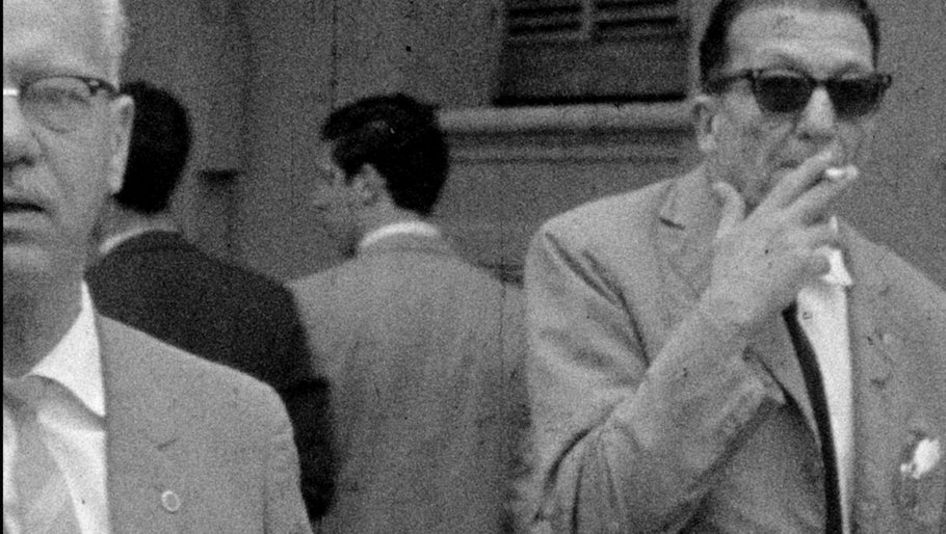 Filmstill aus BAJO LAS BANDERAS, EL SOL: Ein verschwommenes Fernseh-Bild in schwarz-weiß. Man sieht zwei ältere Männer in Anzug auf der Straße, einer raucht eine Zigarette. Im Hintergrund sind weitere Männer.