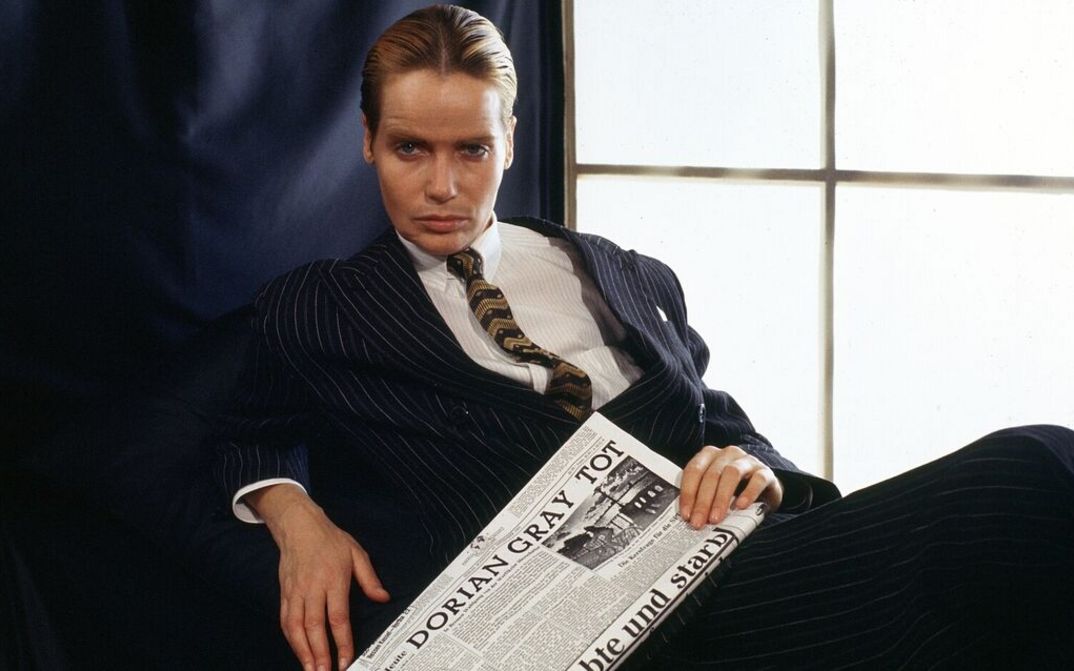 Film still from DORIAN GRAY IM SPIEGEL DER BOULEVARDPRESSE: The actress Veruschka von Lehndorff as Dorian Gray sits in a suit on an armchair and looks into the camera, holding a newspaper in her hand.