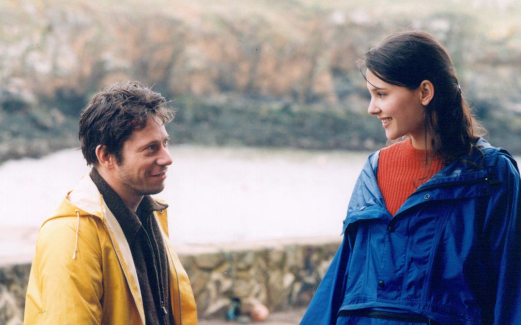 Film still from FIN AOÛT, DÉBUT SEPTEMBRE. A man and a woman in rain jackets smile at each other.