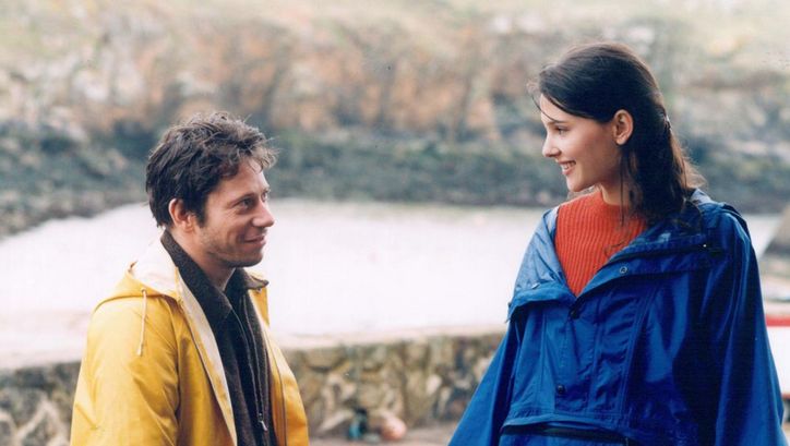 Filmstill aus FIN AOÛT, DÉBUT SEPTEMBRE. Ein Mann und eine Frau in Regenjacken lächeln sich an.