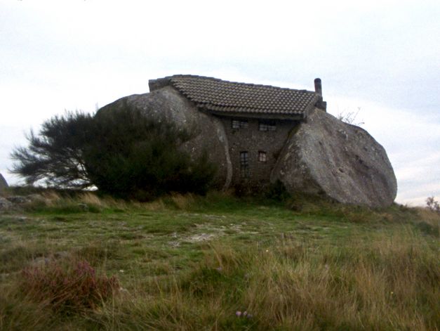 Filmstill from Deborah Stratman’s „Last Things“. A cobblestone house on a meadow built between two erratic boulders. In the background a clouded sky.