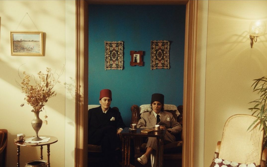 Film still from NEO NAHDA: Two women dressed as men sit between a door frame and look directly into the camera.